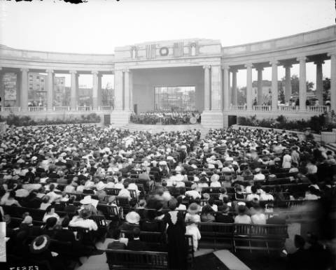 People crowd the plaza at the Greek Theater and the Colonnade of Civic Benefactors at Civic Center in Denver, Colorado; the Denver Municipal Band is on the stage, and folding wooden chairs provide seating for the audience. (circa 1920-1930)