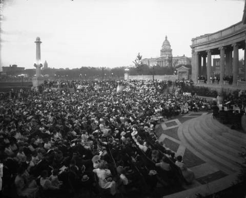 People crowd the plaza at the Greek Theater and the Colonnade of Civic Benefactors at Civic Center in Denver, Colorado; an orchestra is on the stage, and folding wooden chairs provide seating for the audience. The Colorado State Capitol, Presbyterian Church, and YMCA building are in the background. (circa 1920-1930)