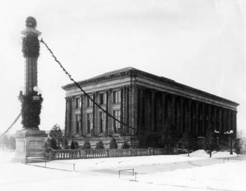 View of the Denver Public Library at Colfax Avenue and Bannock Street in the Civic Center neighborhood of Denver, Colorado. The Greek Revival style building has columns and is decorated with greenery for Christmas. A column is in Civic Center Park nearby. Snow covers the ground. (1920-1930)