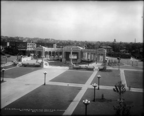 View of the Greek Theater and Colonnade of Civic Benefactors in Civic Center, Denver, Colorado (architects Willis A. Marean and Albert J. Norton; completed in 1919); shows pedestrian traffic in park, automobile traffic on roads behind theater (Broadway, 14th Street, Acoma), businesses in background, Ford O'Meara-Green Motor Company, Fisk Tires, and turret of Evans School in distance. (circa 1920)
