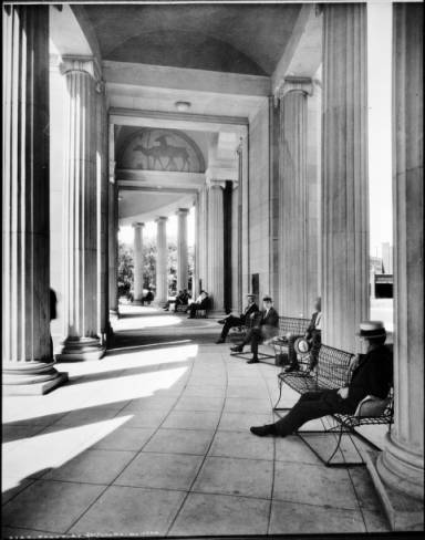 Interior view of the Voorhies Memorial pergola, looking east, in Denver Colorado's Civic Center (designed by Fisher and Fisher); Denver artist, Allen True painted silhouette elk directly on concrete, high on lunette of arch; shows people seated on wire benches under roof of colonnade, tire and motor businesses in distance between columns. (1922-1925)