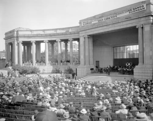 An audience sits in rows of wooden chairs in the Greek Theater and Colonnade of Civic Benefactors, Civic Center Park, Denver, Colorado. An employees' shop band of the Denver & Rio Grande Western Railroad performs on the stage under a banner that reads: "Denver & Rio Grande Western Scenic Line Bands Alamosa, Salt Lake, Brunham." (circa 1920-1930)