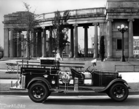 The Idaho Springs, Colorado, fire truck is in front of the Denver  Civic Center. The truck is equipped with an axe, two fire extinguishers, ladders, fire hooks, a small, pressurized water container, a fire bell, a large head lamp mounted where the windshield would be, and a flashlight. There is a two person front seat, and running boards surround the body. "Idaho Springs Fire Dept.," is painted on the hood. Behind the truck, ionic  columns accent Civic Center structures. (6-1-1929)